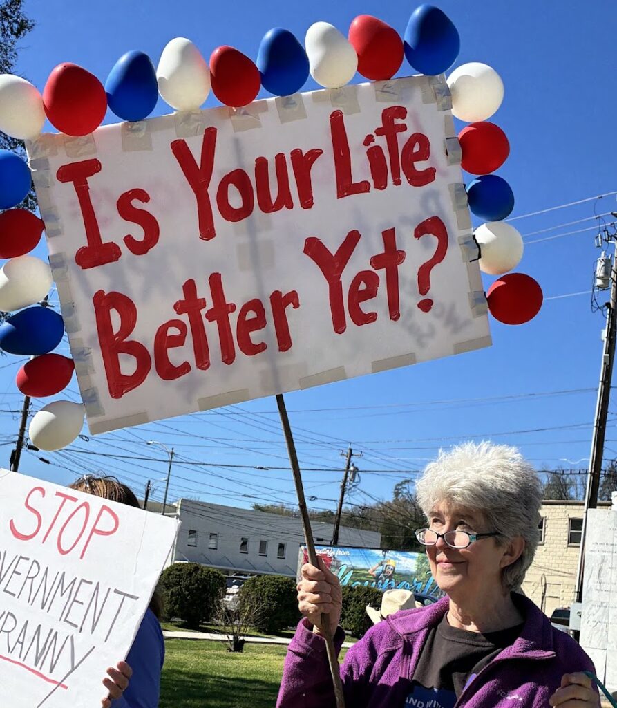 An older woman holding up a protest sign that says, "Is your Life Better Yet?"